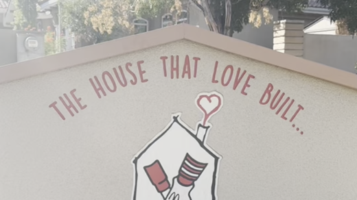 A close-up of a building wall displaying the Ronald McDonald House Charities sign. It features the text: "THE HOUSE THAT LOVE BUILT... RONALD MCDONALD HOUSE CHARITIES & GREATER LAS VEGAS 2323 POTOSI ST." and an emblem of hands holding within a house outlin